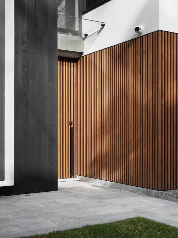 Contemporary residential exterior courtyard or entry area featuring a tall vertical timber slat privacy gate and fence, dark vertical cladding, white rendered walls, a glass balustrade, and grey stone or concrete paving. The composition highlights strong geometric lines, contrasting black, warm wood, white, and grey materials, with a small lawn edge in the foreground.