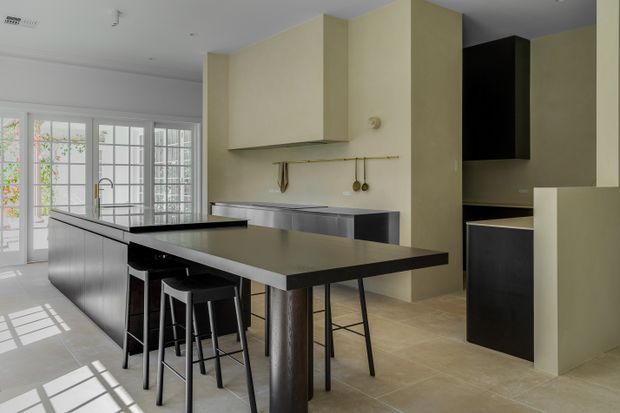 Contemporary kitchen with a large dark island and integrated breakfast bar, black stools, pale painted cabinetry and walls, stainless-steel work surfaces, and light tiled flooring. Full-height glazed French doors and windows bring in abundant natural light, while a tall dark pantry or appliance cabinet creates contrast.