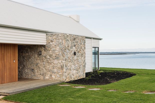 Exterior view of a low, contemporary coastal house beside a broad body of water. The facade combines pale horizontal weatherboard cladding with a prominent irregular natural-stone wall, a light pitched roof, vertical timber entry door, timber deck threshold, and a narrow glazed corner window. Landscaped lawn and garden beds surround the building.