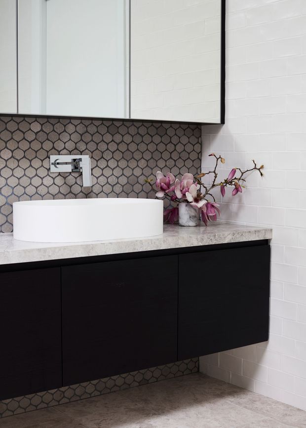 Contemporary bathroom vanity with a white vessel basin on a pale stone benchtop, matte black wall-mounted cabinetry, chrome wall-mounted tapware, and a decorative dark patterned mosaic splashback. White rectangular wall tiles and a large frameless mirror create a clean, high-contrast finish.