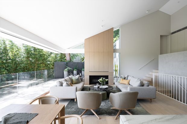 Bright, spacious contemporary open-plan living area with a high sloping ceiling, expansive glazing, and a generous outdoor terrace beyond glass balustrading. The room features a large light-toned sectional sofa, sculptural lounge chairs, a dark round coffee table, a timber dining table, pale timber flooring, and a tall timber-clad fireplace wall. A white-railed stair and upper-level void add architectural interest.