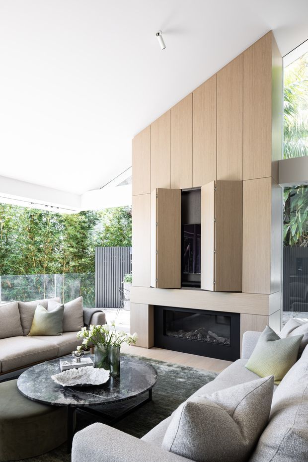 Bright contemporary living room with floor-to-ceiling glazing opening to lush greenery, a large light timber built-in media and storage wall, and a black fireplace set beneath concealed cabinetry. Neutral upholstered sofas, a round dark coffee table, soft cushions, and layered rug create a refined, inviting seating area.