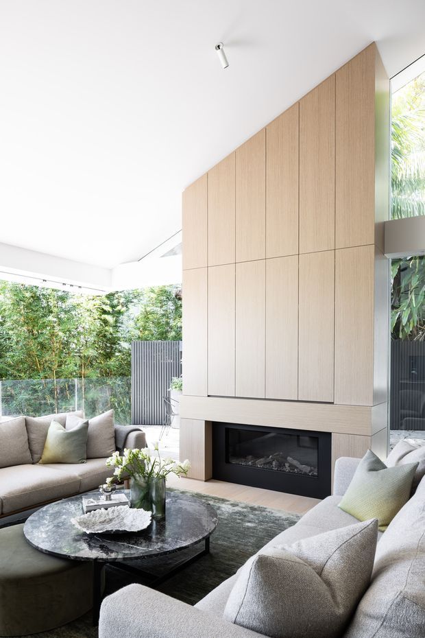 Bright contemporary living room with floor-to-ceiling glazing, lush greenery visible outside, a large built-in timber-clad fireplace wall, and a sophisticated seating arrangement around a dark round coffee table. The room has a high, angled ceiling, neutral upholstered sofas, a textured rug, and abundant natural light.