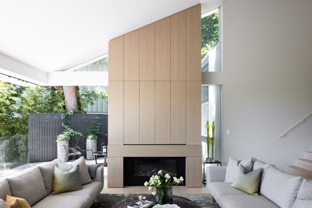Bright contemporary living room with two light upholstered sofas facing a central fireplace clad in warm vertical timber cabinetry. Floor-to-ceiling glazing opens to a leafy outdoor setting, while a staircase rises along the right wall. The room has a clean, refined palette of soft grey, white, and light timber with abundant natural light.