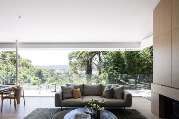 A refined contemporary living room with a grey upholstered sofa, coffee table, dining area, and extensive floor-to-ceiling glazing opening onto a leafy landscape. The room features pale timber built-in cabinetry, a minimalist fireplace wall, a smooth white ceiling, and a neutral, softly lit palette.