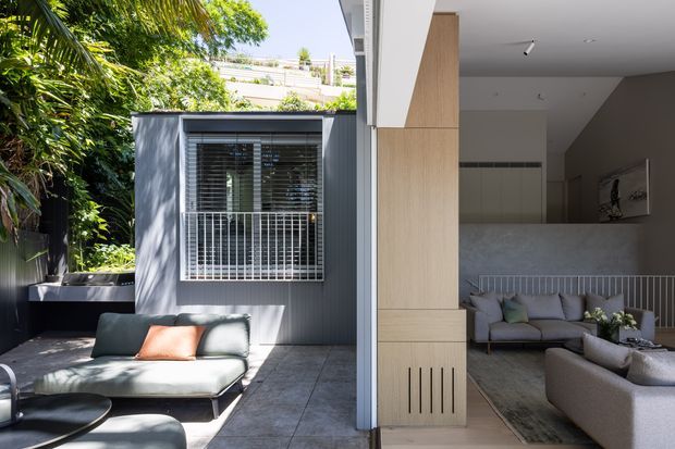 A contemporary indoor-outdoor living space with a covered patio or terrace opening directly into a refined living room. The exterior features dark vertical cladding, lush planting, tiled paving, and a large window with security bars and horizontal blinds. Inside, a neutral-toned living area includes sofas, a rug, a sculptural wall artwork, a pale stone or concrete fireplace wall, and built-in light timber cabinetry dividing the spaces.