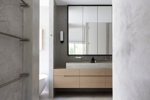 Contemporary bathroom viewed through a doorway, featuring a floating timber vanity with a pale stone benchtop, integrated basin, black tapware, large mirrored cabinet, dark small-format tiled backsplash, and textured grey plaster walls. A partially visible toilet and heated towel rails appear on the left.