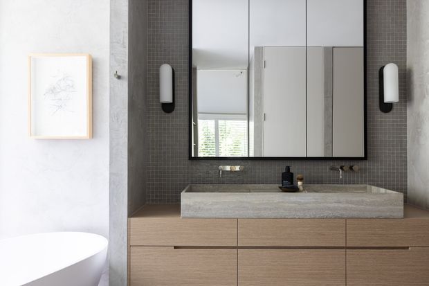 Contemporary bathroom vanity with a long floating timber-toned cabinet, integrated grey stone basin, dark small-format tiled splashback, mirrored wall cabinet, black wall lights and a partially visible freestanding white bathtub. The space has a refined, minimalist, neutral palette with contrasting dark tile.