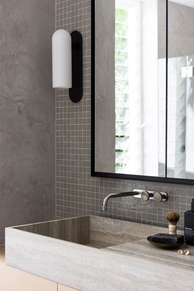 Close-up of a refined contemporary bathroom vanity featuring a large black-framed mirror, wall-mounted brushed metal tapware, a grey stone or concrete benchtop with integrated basin, grey mosaic tile splashback, textured grey wall finish, and a minimalist white wall sconce. Personal grooming products sit on the vanity.