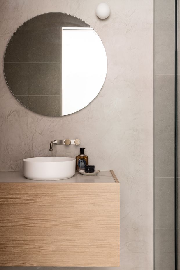 A refined contemporary bathroom vanity with a round white vessel basin, wall-mounted brushed metal tapware, pale timber-grain floating cabinetry, and a large circular mirror. The walls are finished in a warm grey-beige textured surface, with dark grey tiled wall visible at the right edge and a small spherical wall light above the mirror. The composition is minimalist and softly lit.