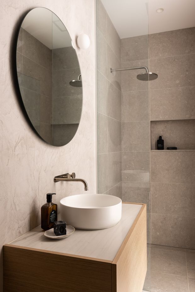 A compact contemporary bathroom featuring a floating timber vanity with a white vessel basin and wall-mounted brushed metal tapware. An oval black-framed mirror hangs above the basin, while a frameless glass shower enclosure with a round rain showerhead occupies the right side. Large grey-beige stone-look tiles line the shower walls and floor, with a recessed shower niche and subtle warm lighting.