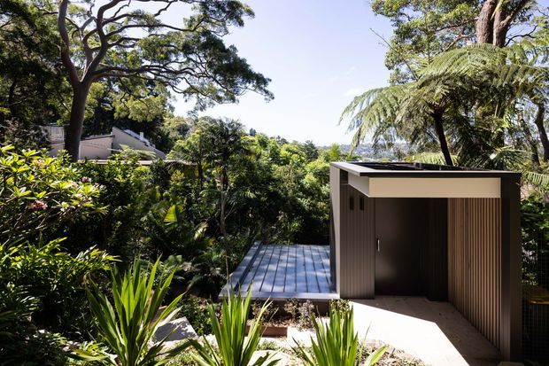 A compact contemporary garden structure or outbuilding set within dense subtropical landscaping. The building has a flat roof, dark vertical ribbed cladding, a recessed opening, and a narrow timber-toned deck or walkway leading through the vegetation. Mature trees and lush planting surround the structure.