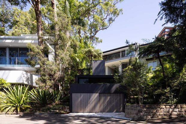 Street-facing view of a contemporary residential property nestled among mature trees and tropical landscaping. The composition shows a dark, vertically ribbed garage door and matching privacy gate, pale rendered walls, extensive glazing, balconies, and a low stone retaining wall. The multi-level building has a low-pitched roofline and is integrated into a leafy suburban setting.