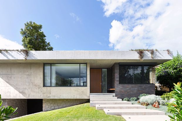 Modern single-storey residence with a strong horizontal composition, flat concrete roof slab, exposed concrete walls, dark brickwork, large black-framed glazing, and a timber entry door. Broad stepping-stone paths cross a manicured lawn, while trailing planting softens the roof edge. The image is photographed from the front exterior under a bright blue sky.
