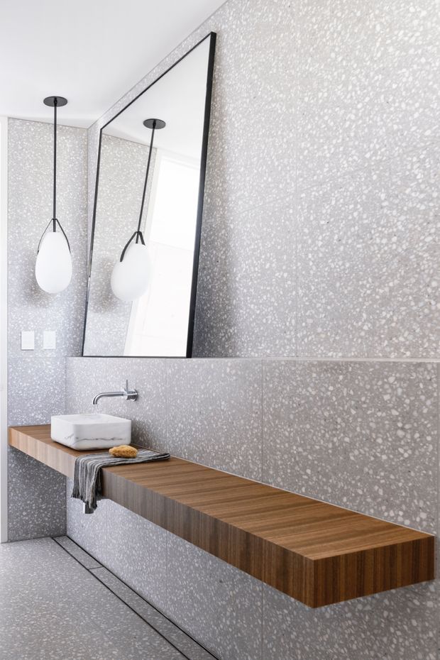 Contemporary bathroom featuring grey terrazzo wall and floor surfaces, a large frameless-style rectangular mirror with dark trim, a floating timber vanity bench, and a white vessel basin with wall-mounted chrome tapware. A sculptural pendant light hangs beside the mirror, creating a refined, textured monochromatic scheme with warm timber contrast.