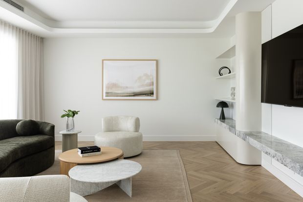 A bright, refined living room with a neutral palette and layered seating arranged around a pair of round coffee tables. The room features white built-in media cabinetry and shelving, a large wall-mounted television, a pale stone-look benchtop or ledge, herringbone timber flooring, sheer curtains, recessed cove ceiling detailing, and understated contemporary furnishings.