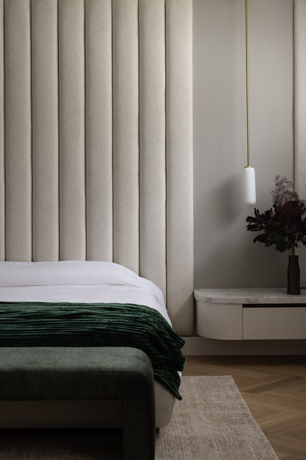 A refined bedroom detail featuring a tall, vertically channelled upholstered headboard in warm off-white fabric, crisp white bedding, a deep forest-green throw, and a dark upholstered bed base. A curved floating bedside cabinet with a pale stone top sits beside the bed, paired with a slim pendant light and dark floral arrangement. Herringbone timber flooring and a neutral textured rug complete the calm, luxurious composition.
