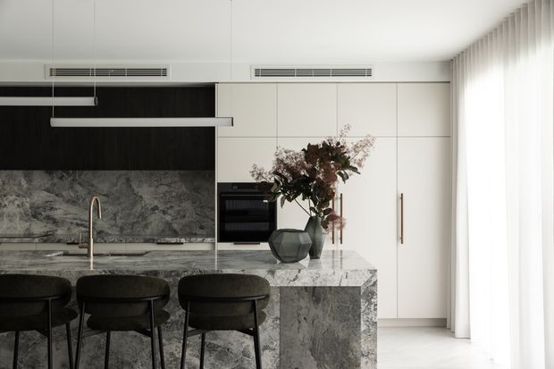 Contemporary kitchen with a large grey veined stone island and matching splashback, dark timber-toned cabinetry, tall white built-in cabinetry, and three dark upholstered bar stools. A linear pendant light hangs above the island, with sheer full-height curtains along the right side and soft natural light filling the space.