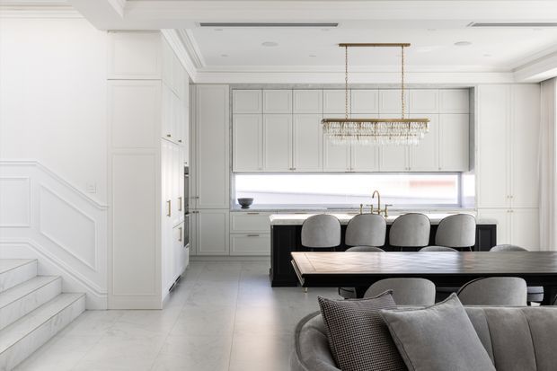 Bright, refined open-plan kitchen and dining area in a predominantly white and light grey interior. The kitchen features extensive panelled cabinetry, a long horizontal window, pale stone-look flooring, and a dark island or dining counter. A large rectangular crystal chandelier hangs above the dining table, with a curved sectional sofa in the foreground and a staircase with decorative wall panelling at left.