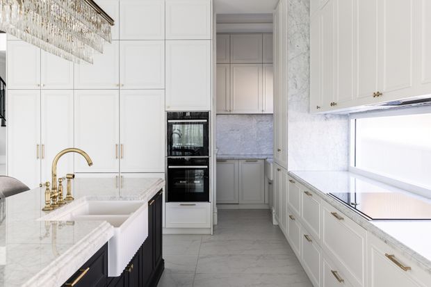 Bright, refined galley-style kitchen with extensive white raised-panel cabinetry, a dark island base, white marble-look benchtops and splashbacks, brass hardware and tapware, integrated black appliances, and a large linear crystal chandelier. A pantry or secondary kitchen area is visible through the opening at the rear.