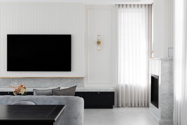 Bright, refined contemporary living room with a wall-mounted television, grey sectional sofa, black low-profile cabinetry, marble-look fireplace surround, pale stone or tile flooring, fluted wall panelling, and full-height sheer curtains. The room has a clean monochromatic palette with black accents and light grey finishes.