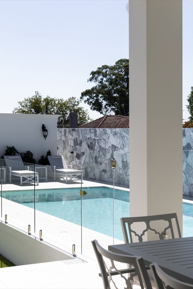 Bright outdoor pool area viewed from a covered patio or terrace. A turquoise rectangular swimming pool is enclosed by frameless glass safety fencing, with white paving, white lounge chairs, dark planted areas, and a striking grey-and-white stone feature wall. A dining table and chairs occupy the foreground beneath tall white structural columns. Nearby tiled residential rooftops and mature trees are visible beyond the boundary wall.