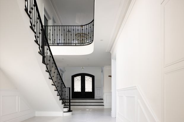 Bright, refined residential entry hall viewed toward a black double front door, with a sweeping curved staircase and ornate black metal balustrade leading to an upper landing. White walls, decorative wall panelling, crisp moulded cornices, pale flooring, and recessed lighting create a high-contrast, formal interior.