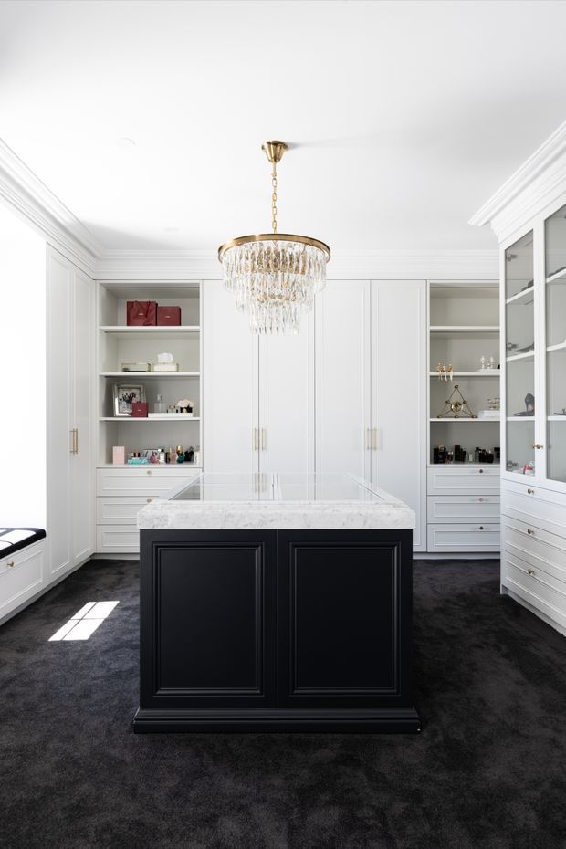 A bright, refined walk-in wardrobe with extensive white built-in cabinetry, glass-front display cabinets, open shelving and drawers surrounding a central black island with a white stone benchtop. A crystal chandelier hangs from the white decorative ceiling, while dark plush carpet creates contrast with the light joinery and brass hardware.