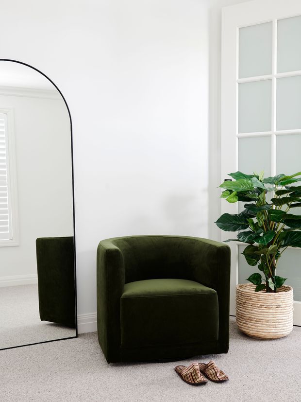 Bright, minimalist interior corner featuring a deep olive-green upholstered barrel chair, a large black-framed arched floor mirror, a potted leafy plant in a woven basket, white walls and window trim, and a light neutral carpet. The mirror reflects the chair and nearby shuttered window, while slippers rest on the floor in front.