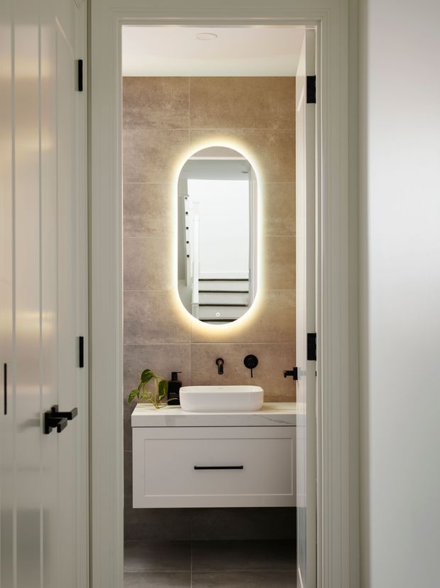 A compact contemporary bathroom viewed through an open doorway. The room features a floating white vanity with a vessel basin, black tapware and handles, large warm taupe stone-look wall tiles, dark floor tiles, and an illuminated oval mirror. White paneled doors and trim frame the bathroom entrance.