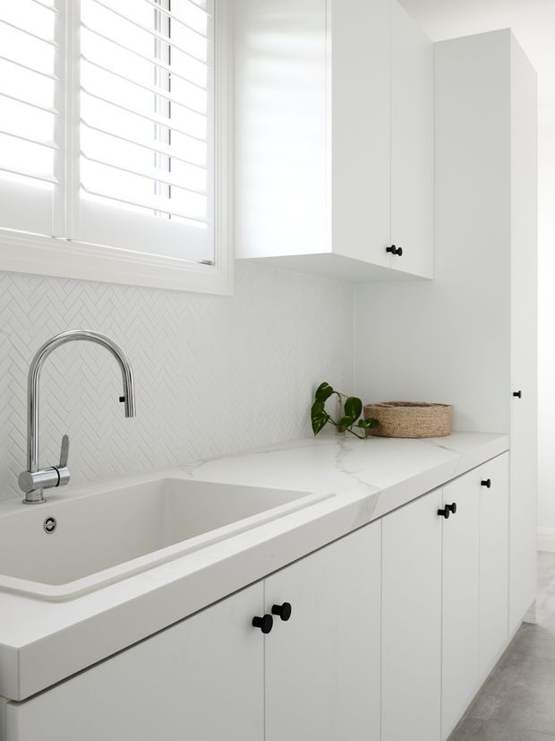 Bright, compact white kitchen or utility area with shaker-style cabinetry, black knobs, a white stone benchtop, integrated sink and chrome gooseneck tap. A white herringbone tile splashback sits beneath plantation shutters, with a small plant and woven basket adding subtle texture.