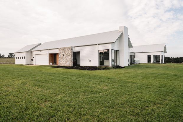 A low, single-storey residential building set within a broad mown lawn. The white exterior combines horizontal weatherboard-style cladding, pale metal roofing, dark-framed glazing, and a prominent irregular natural-stone wall around the entry and garage area. Several connected wings create a simple contemporary rural homestead form, with a largely open landscape visible around it.