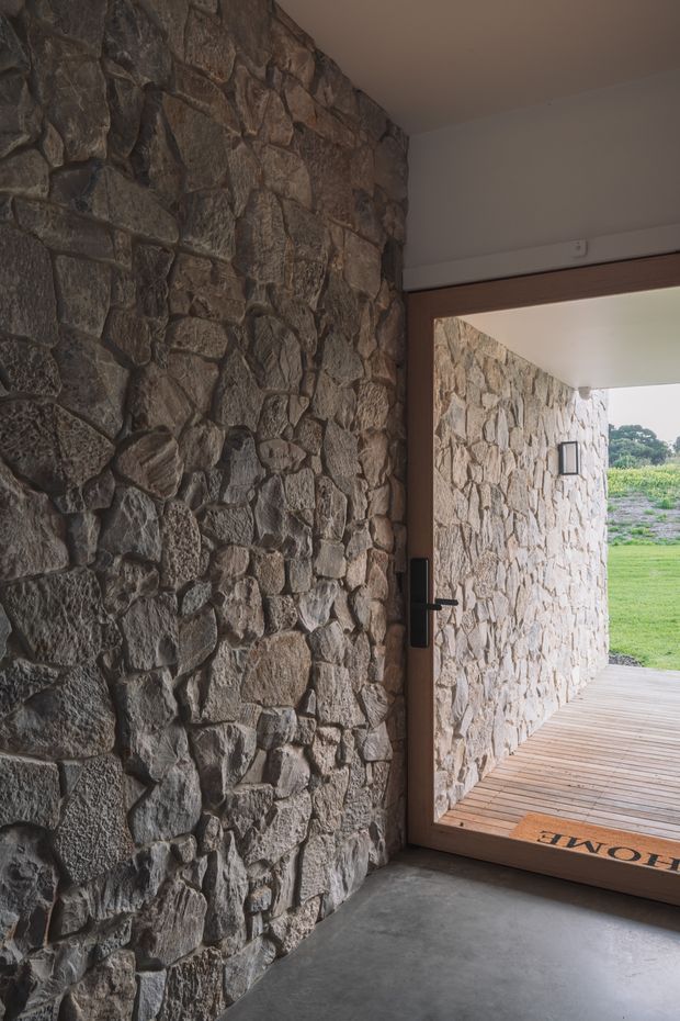 A rustic entryway with exposed irregular natural-stone walls, a smooth concrete floor, and a large timber-framed glazed door opening onto a timber deck and grassy landscape. The stone continues onto the exterior wall visible through the doorway, creating a strong indoor-outdoor connection. The space is dim and earthy, with a simple black door handle and minimal detailing.