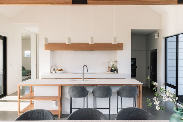 Bright contemporary kitchen with a large white island and three upholstered bar stools, set beneath a timber ceiling beam and matching open shelf. The kitchen features white cabinetry, a pale stone island benchtop, minimalist pendant lights, dark-framed glazing, and a view toward an adjacent living or bedroom area.