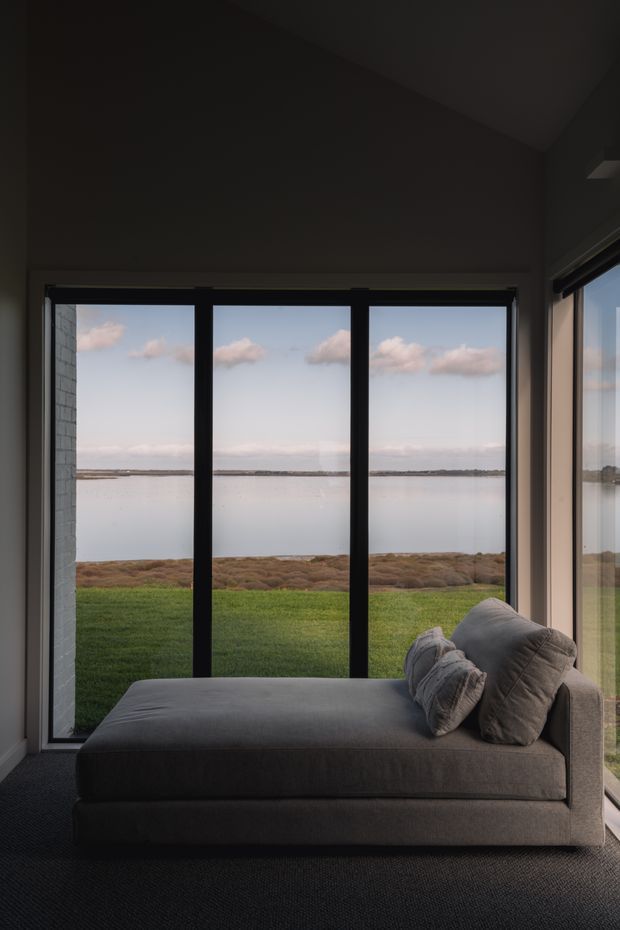 A dark, calm interior corner with a large upholstered chaise lounge positioned in front of full-height black-framed glazing. The room overlooks a tranquil body of water and grassy shoreline, creating a strong indoor-outdoor connection. The space has charcoal carpeting, dark walls, minimalist detailing, and expansive floor-to-ceiling windows.