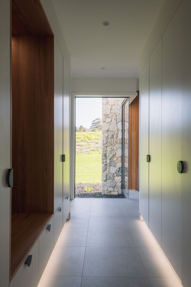 A narrow, refined hallway or entry passage lined with full-height white built-in cabinetry on both sides. The left side features a warm dark timber-lined open bench niche and drawers, while concealed linear lighting illuminates the pale large-format tiled floor. The passage leads to a glazed exterior doorway, with a natural stone wall and landscaped grounds visible beyond. The ceiling is smooth with recessed downlights, and dark hardware provides subtle contrast.