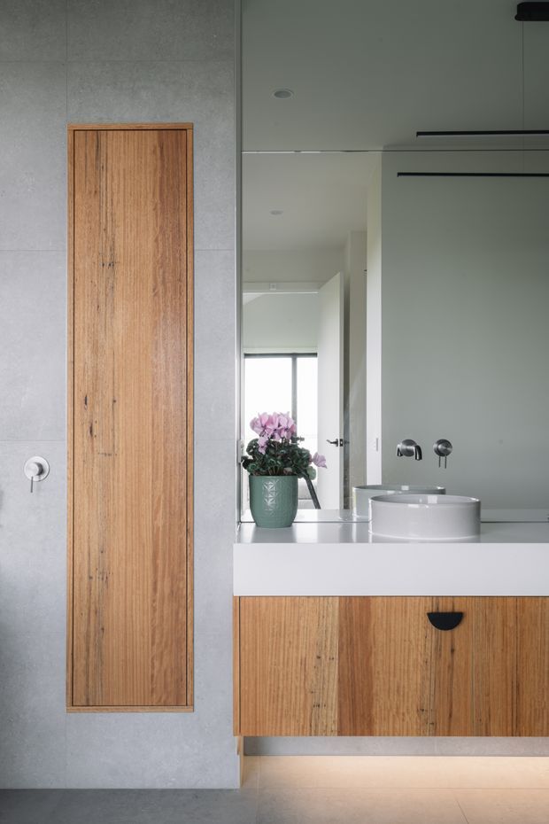 A refined contemporary bathroom vanity with a floating timber drawer unit, white solid-surface benchtop, and a round vessel basin. The space features a large wall mirror, pale grey wall surfaces, a tall timber-panelled storage cabinet, minimalist black linear lighting, and concealed under-vanity illumination. A small potted flower provides a subtle decorative accent.