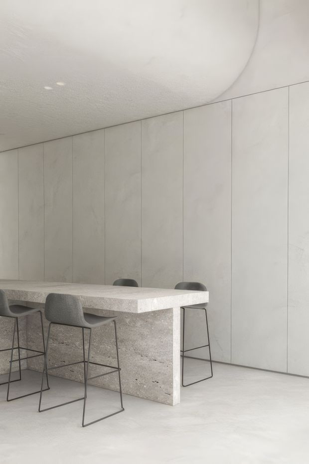 A minimalist monochromatic interior featuring a large pale concrete or stone island with an integrated thick benchtop and waterfall-style side, surrounded by dark-framed grey bar stools. Full-height vertically panelled light walls and a softly textured curved ceiling create a seamless, sculptural space.