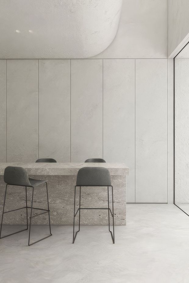 Minimalist dining area with a long stone island or table and four dark upholstered bar stools. The space features pale large-format wall panels, a smooth light concrete-like floor, and a rounded sculptural ceiling element. Full-height glazing or a glass door is visible at right, bringing in soft natural light. The restrained grey-beige palette and simple geometric forms create a calm, refined interior.