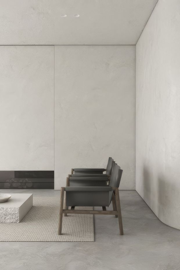 Minimalist living room with a row of sculptural dark grey lounge chairs arranged along a textured neutral rug. The space features pale grey rendered walls and ceiling, a smooth grey concrete-like floor, a low white textured coffee table, and a dark linear fireplace or built-in feature along the rear wall. Recessed ceiling lights and a curved wall edge create a restrained, gallery-like atmosphere.