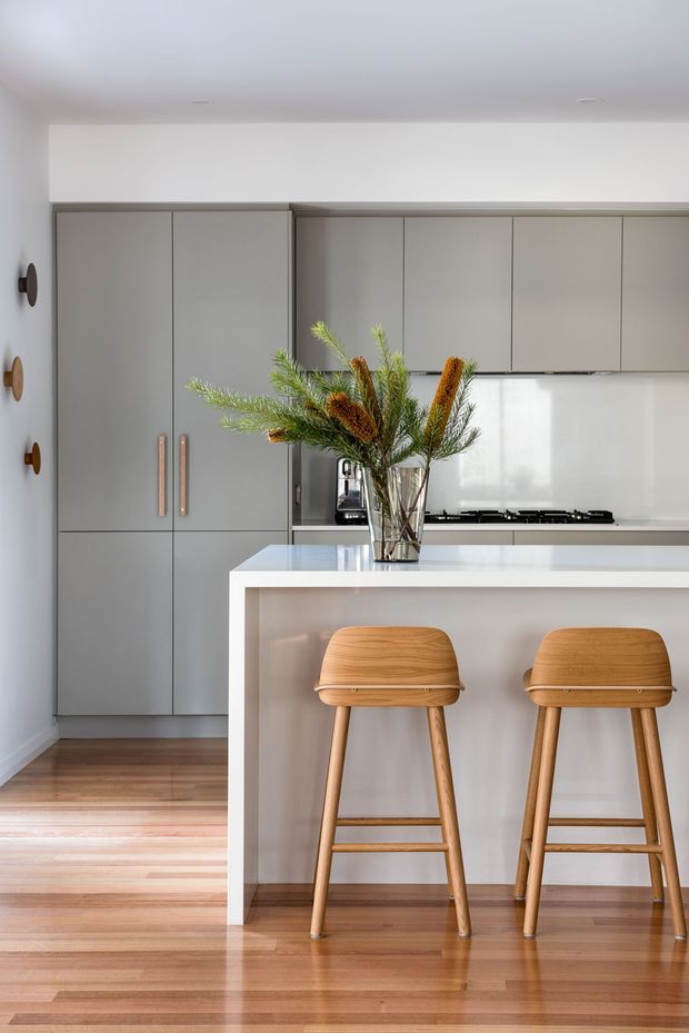 A bright, compact contemporary kitchen with a white island and integrated seating, matte grey floor-to-ceiling cabinetry, white splashback, timber flooring, and warm timber bar stools. A glass vase with greenery provides a natural centrepiece, while slim brass-toned handles and wall-mounted accents add subtle detail.