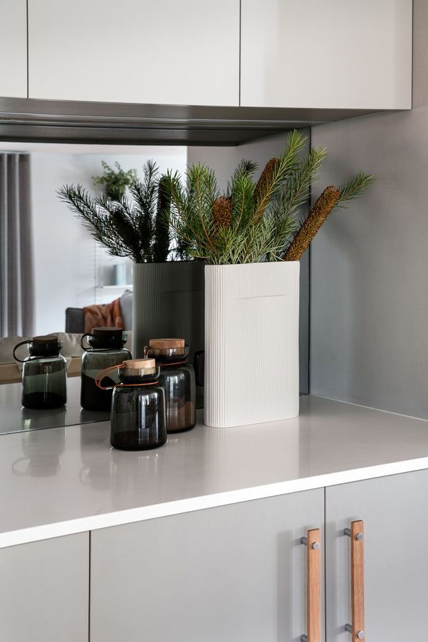 Close-up view of a contemporary kitchen benchtop with pale grey cabinetry, a smooth light benchtop, dark glass storage jars, and tall ribbed ceramic planters containing greenery. A mirrored splashback reflects the adjacent dining or living area. The cabinetry has long timber-and-metal handles, and the overall composition is refined and minimalist.