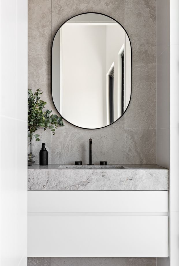 A refined minimalist bathroom vanity with a floating white drawer cabinet, grey stone-look wall tiles, and a matching stone benchtop. Matte black tapware and an oval arched mirror provide contrast, while a small leafy plant adds a natural touch.