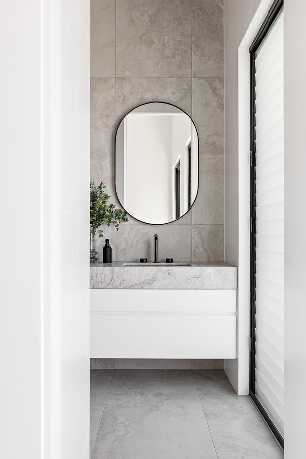A narrow, bright contemporary bathroom viewed through a doorway, featuring a floating white vanity with a grey marble-look stone benchtop, black tapware, a rounded black-framed mirror, large grey stone-look wall and floor tiles, and a white louvred window covering.