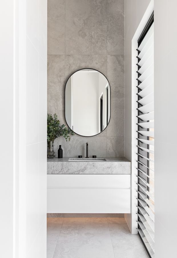 A compact contemporary bathroom vanity area with a floating white cabinet, grey stone-look tiled wall and benchtop, black mixer tap and oval black-framed mirror. White plantation shutters provide filtered daylight along the right side, while a small plant and dark bathroom accessories add contrast.
