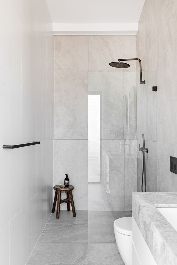 Contemporary minimalist bathroom with a walk-in glass shower, black rain shower and fittings, large light grey stone-look wall and floor tiles, a white toilet, and a pale stone vanity with an integrated basin. A narrow vertical window provides natural light, while a small wooden stool adds warmth.