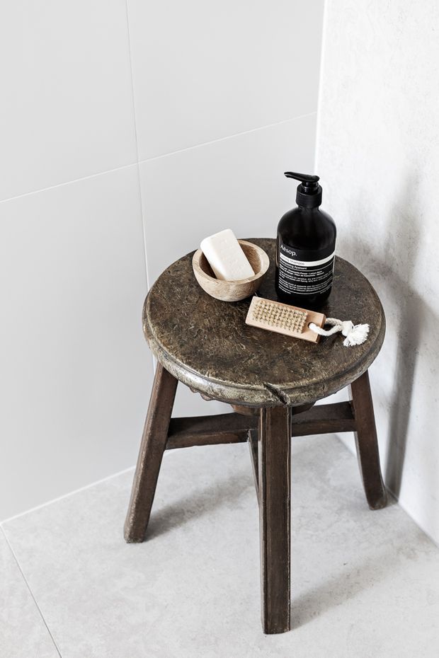 A close-up vignette in a minimalist bathroom, featuring a rustic dark wooden stool with a worn round stone-like top. The stool holds a black pump soap dispenser, a small wooden bowl with a white soap bar, and a natural-bristle body brush. Pale large-format wall and floor tiles and a textured white wall create a neutral, understated setting.