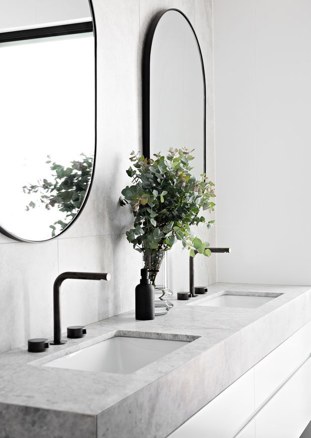 Contemporary double-vanity bathroom featuring a long pale stone countertop with integrated rectangular basins, matte black wall-mounted tapware and slim black-framed arched mirrors. White tiled walls and white built-in cabinetry create a clean, minimal scheme, with a leafy arrangement adding a natural accent.