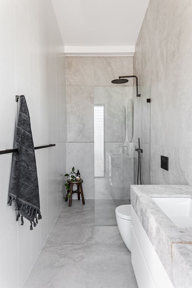 A narrow, contemporary bathroom with a walk-in glass shower, freestanding-style white bathtub, wall-mounted shower fittings, and a white toilet. Large pale grey marble-look tiles cover the floor and walls, while a dark towel rail, black fixtures, and a small plant provide contrast. The space is bright and minimalist with a high white ceiling.