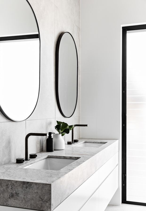 A bright contemporary bathroom vanity featuring a double rectangular inset basin in a thick pale grey stone benchtop, white built-in cabinetry, matte black tapware, and large rounded-rectangle mirrors with black frames. A black-framed glazed door or window with white horizontal blinds is visible at right, while a small plant and monochrome vessels add restrained styling.